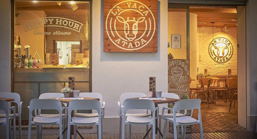 Terraza de un restaurante. Se observa la fachada del restaurante y dos mesas de color blanco con cuatro sillas cada una, también de color blanco